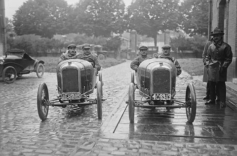 Two small cyclecars facing the camera on wet cobbles, two men in each, a man in a long coat beside them. Opens a larger view