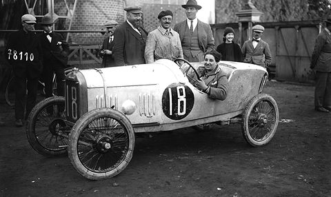 A smiling woman at the wheel of a white cyclecar numbered 18, men in caps and a woman standing behind. Opens a larger view