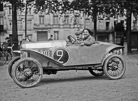 A woman and a man in a small pale cyclecar numbered 2 on a dirt square, town houses and trees behind. Opens a larger view