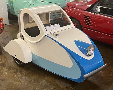 A teardrop-shaped blue and white plywood three-wheeler with a single headlamp in its nose and an arched open cabin. Opens a larger view
