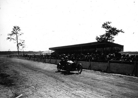 A small dark cyclecar with two aboard racing along a dirt road past a fenced, roofed grandstand packed with spectators. Opens a larger view