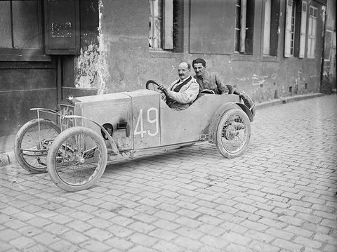 Two men in a small pale cyclecar numbered 49 on a cobbled street before an old plastered building with shutters. Opens a larger view