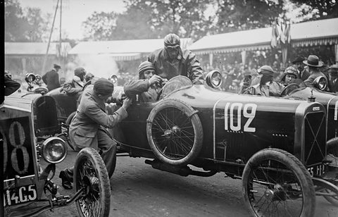 Men crowd around a dark racing cyclecar numbered 102, one leaning in to a driver in a leather helmet. Opens a larger view
