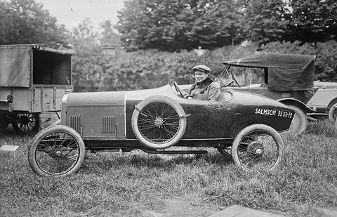 Side view of a dark cyclecar with a spare wheel on its flank and Salmson on the tail, on grass by lorries. Opens a larger view