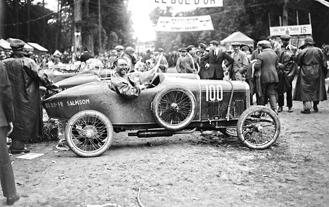 A grinning driver with a bouquet in a muddy racing cyclecar numbered 100, a crowd and a banner reading Le Bol d’Or behind. Opens a larger view