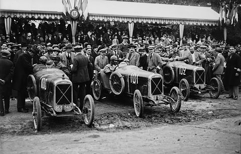 Three racing cyclecars numbered 100, 101 and 102 before a dense crowd under a striped awning. Opens a larger view