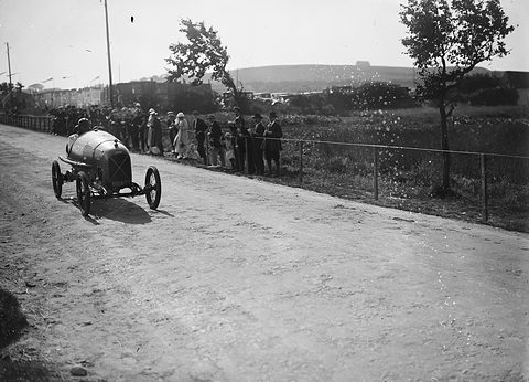 A small racing cyclecar on a pale country road lined by spectators, a fence and windblown trees beside it. Opens a larger view