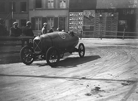 A racing cyclecar with a pointed tail rounds a dirt bend past spectators and a wall of oil advertisements. Opens a larger view