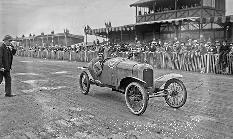 A small racing cyclecar on a dirt track before a wooden grandstand and a crowd behind a fence, a man walking at left. Opens a larger view
