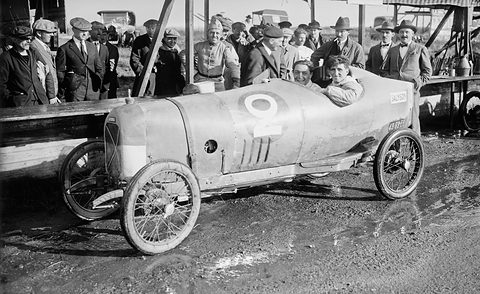 Two men in a pale pointed-tail racing cyclecar numbered 9 on a muddy apron, a crowd at a railing behind.