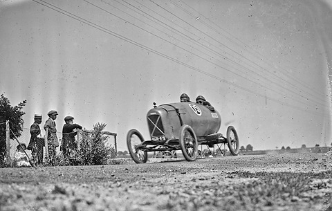 A low pointed-tail racing cyclecar with two crew in helmets on a country road, three men watching from behind a wire fence. Opens a larger view