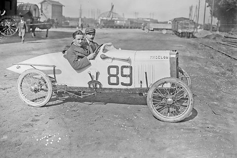 Two men in caps in a pale pointed-tail cyclecar numbered 89 on a dusty yard, railway wagons and sheds behind.