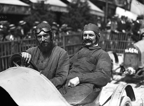 Two men in leather caps and goggles sit side by side in a pale open car, a crowd in straw hats behind a fence.