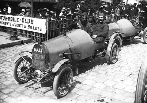 A small open car with a rounded bonnet on cobbles, two men in it, a second car behind and a crowd at a railing with an Automobile-Club sign. Opens a larger view