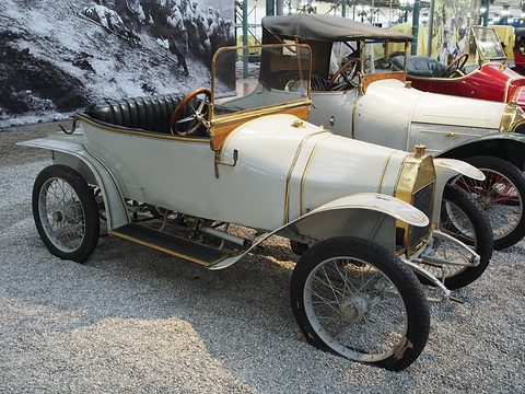 Side view of a grey open two-seater with a brass-trimmed bonnet, black seat and folded hood, on gravel. Opens a larger view