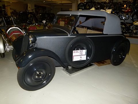 Black small cabriolet with a grey hood and a side-mounted spare wheel, seen from the left front under flash. Opens a larger view