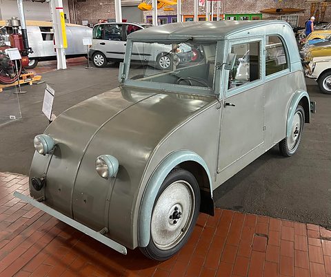 Grey two-door small saloon with a smooth rounded nose, round lamps and an upright cabin on a brick floor. Opens a larger view