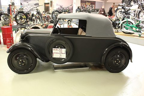 Black small two-door cabriolet with a grey hood up and a spare wheel on its flank, motorcycles behind. Opens a larger view