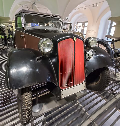 Front of a small saloon: red dummy radiator grille, big headlamps on black wings and a narrow bumper. Opens a larger view