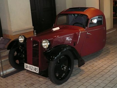 Maroon three-wheeled small car with a closed roof, black wings and a slim grille, on a tiled museum floor.
