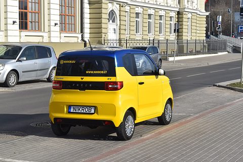 Rear of a yellow boxy city car with a blue roof and a NIKROB plate, parked on brick paving by a yellow building. Opens a larger view