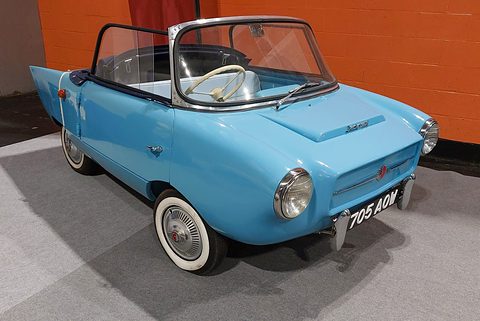 Pale blue open two-seater with whitewall tyres on a grey carpet, an orange wall behind. Opens a larger view