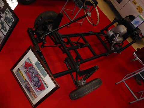 Black tubular frame with wheels, engine and steering column on a red carpet beside framed drawings