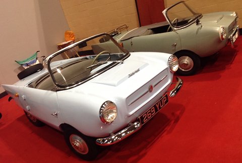 White and pale green open two-seaters on a red carpet in a show stand. Opens a larger view