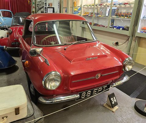 Red small coupé with a chrome grille on a museum floor, a vintage suitcase beside it