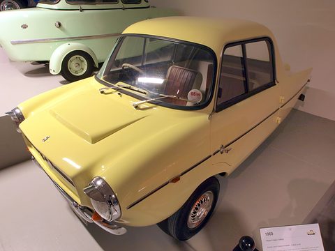 Pale yellow small coupé on a grey museum floor, a green microcar behind. Opens a larger view