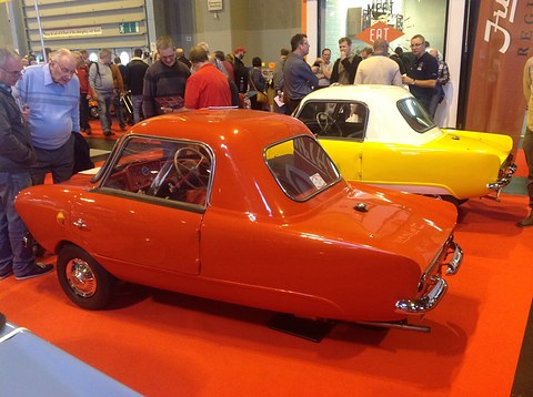 Orange small coupé from the rear left on a red carpet, a yellow and white car and visitors behind. Opens a larger view