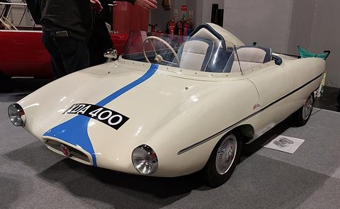 A low white open sports car with a blue stripe over its bonnet, faired headlamps and a wrap-round screen. Opens a larger view