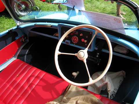 White two-spoke steering wheel over a pale blue dashboard and red seats. Opens a larger view