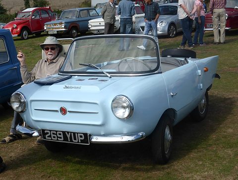 Pale blue open two-seater on grass seen from the front, an older man seated in a chair beside it. Opens a larger view