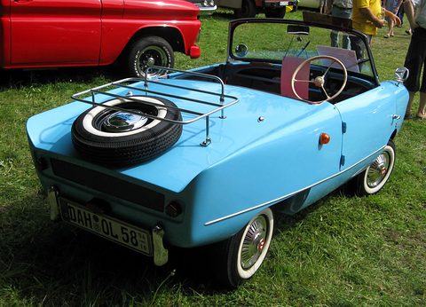 Pale blue open two-seater from behind with a spare wheel on a chrome rack, on grass. Opens a larger view