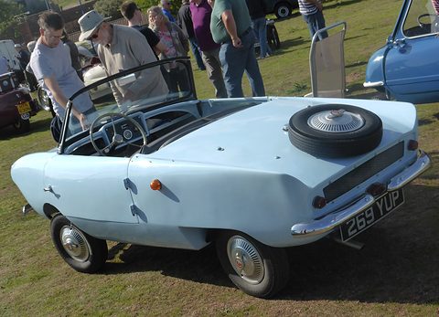 Pale blue open two-seater on grass from the rear right with a spare wheel on its tail, visitors behind. Opens a larger view