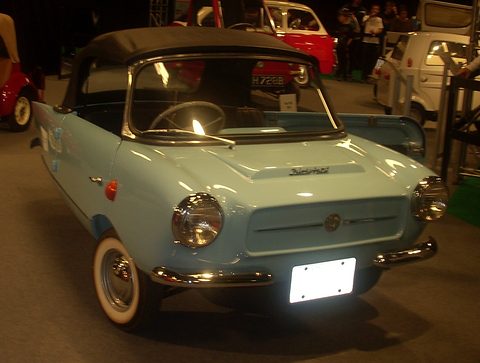 Pale blue two-seater with a black hood and whitewall tyres under dim show lights. Opens a larger view