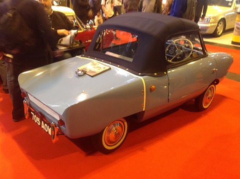Grey two-seater with a navy fabric hood on an orange carpet, seen from behind. Opens a larger view