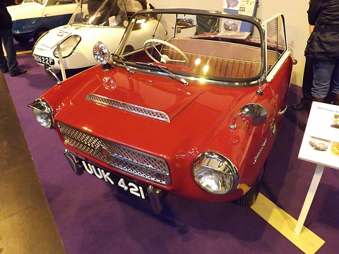 Red open two-seater with a wide chrome mesh grille and round headlamps, seen from the front left. Opens a larger view