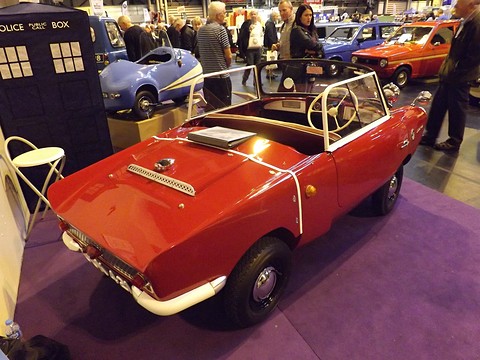 Red open two-seater on a purple show carpet, seen from the rear right. Opens a larger view