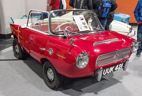 Red open two-seater with a white interior on a grey carpet, seen from the front right. Opens a larger view