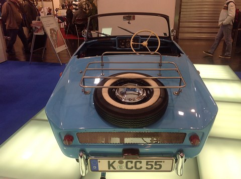 Rear of a pale blue open two-seater with a spare wheel on a chrome rack, on a lit floor. Opens a larger view