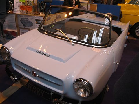 White open two-seater on a dark carpet, seen from the front left. Opens a larger view