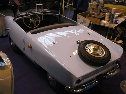 White open two-seater from the rear left with a spare wheel on its tail. Opens a larger view