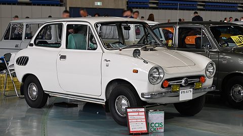 White small saloon with round headlamps and a display board on the floor, visitors behind. Opens a larger view