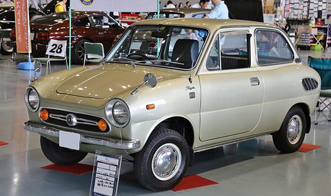Pale gold-green small saloon on a red mat in a sports hall, a display card at its front wheel. Opens a larger view