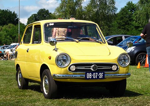 Yellow small saloon on grass seen from the front, a crowd and cars behind. Opens a larger view