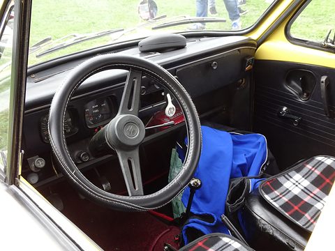 Inside a small car: a three-spoke steering wheel, round dials and a blue bag on a checked seat cover. Opens a larger view