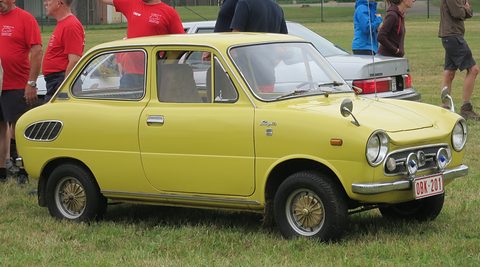 Yellow small saloon on grass with fog lamps and a Belgian plate, people in red shirts behind. Opens a larger view