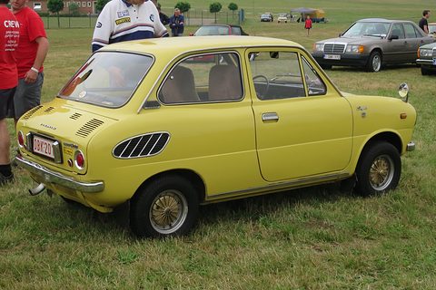 Rear three-quarter of a yellow small saloon on grass with slotted vents and a Belgian plate. Opens a larger view